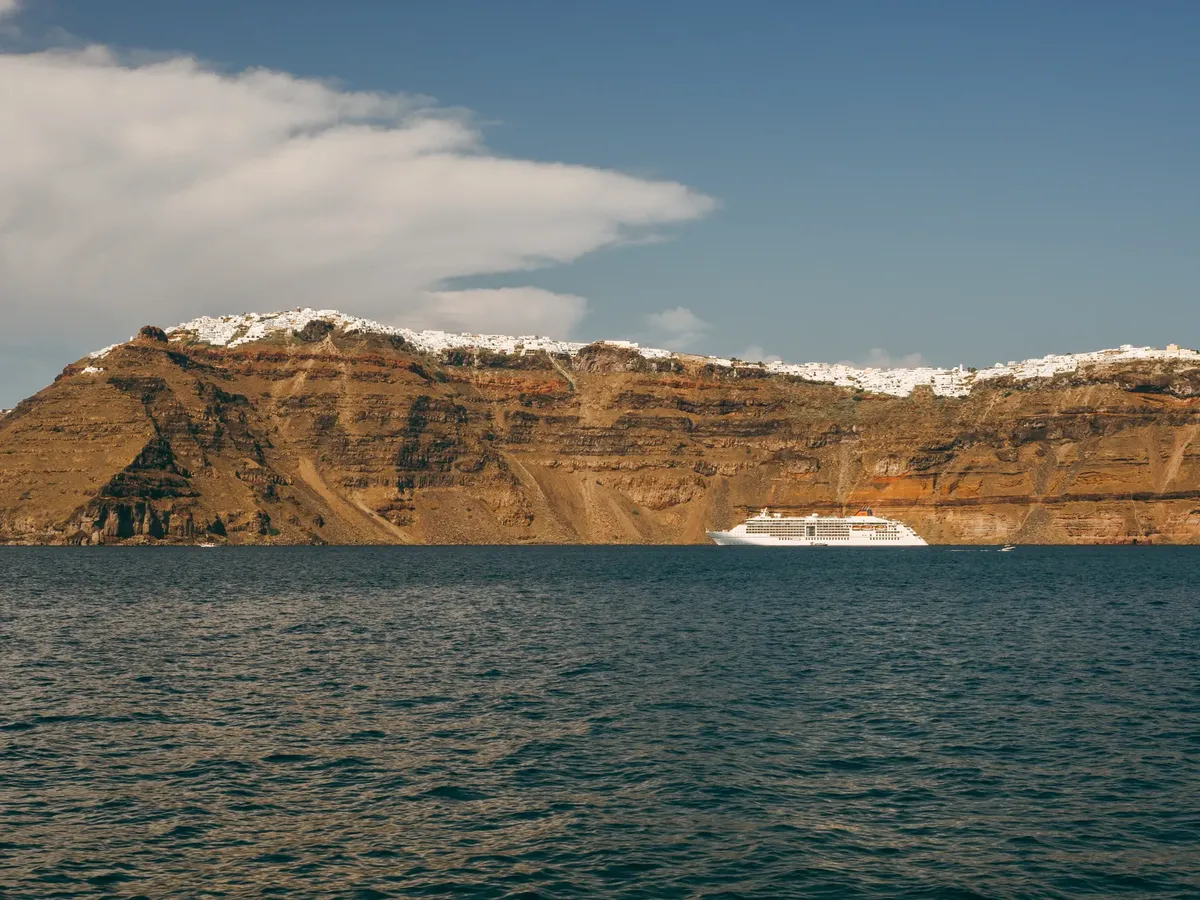 A cruise ship sails by the rugged cliffs of Santorini under a clear sky, capturing the island's serene beauty.