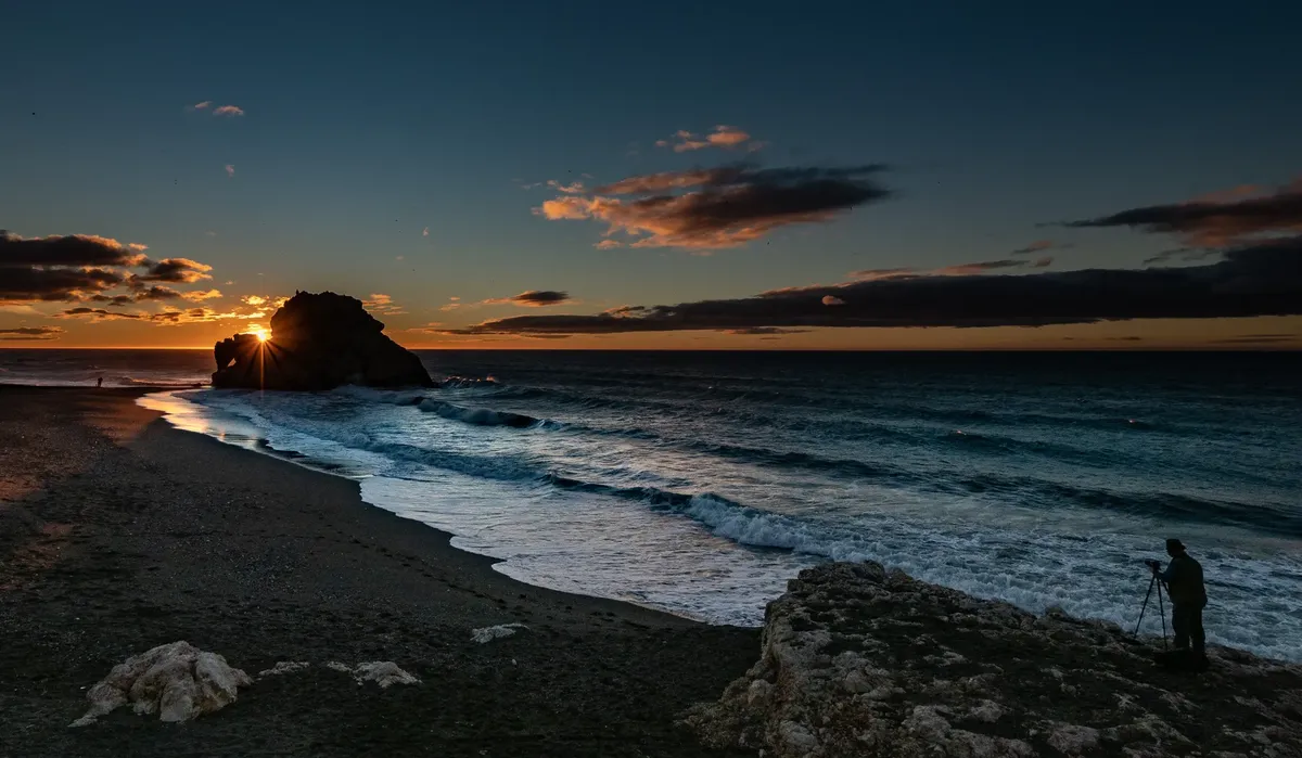 Photographer capturing a stunning sunset at Málaga's scenic coastline with waves and rock formations.
